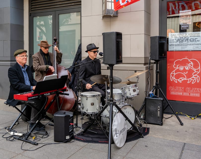 Three musicians play jazz instruments, including a keyboard, upright bass, and drums, outside Cajun Claws.