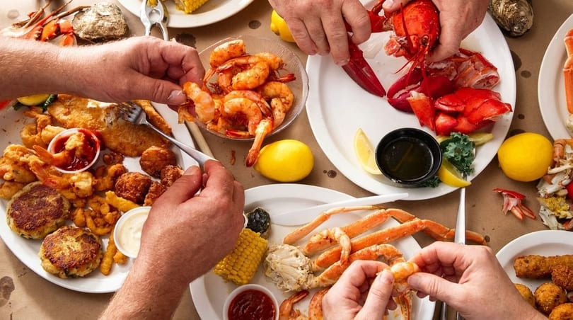 Hands reaching for various seafood dishes, including lobster, crab, shrimp, and fried fish.