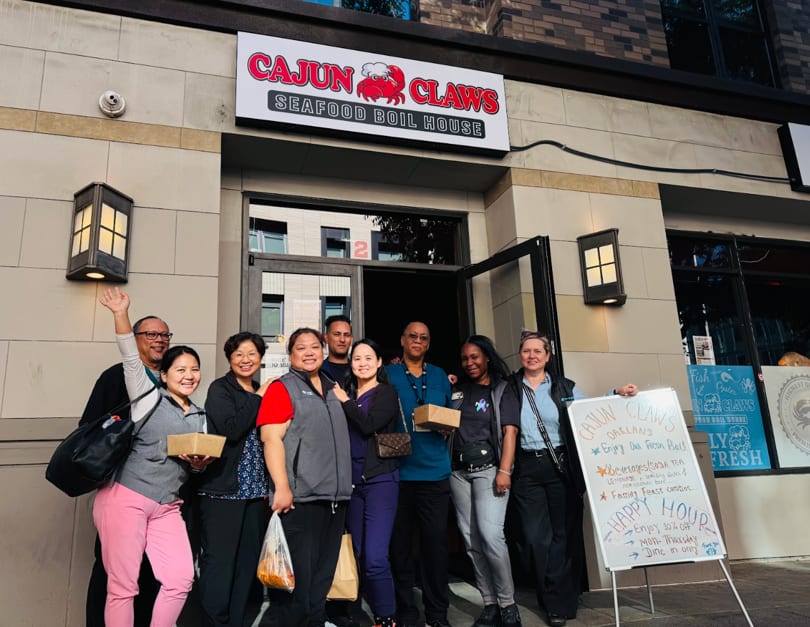 A group of diverse people smiling and holding food containers in front of Cajun Claws Seafood Boil House.
