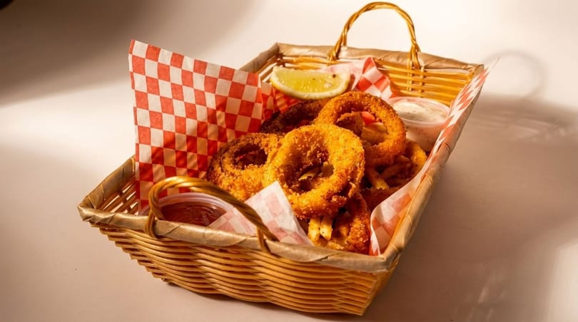A wicker basket filled with golden onion rings, french fries, lemon, and dipping sauces.