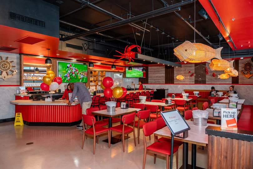 Spacious restaurant interior with red chairs, tables, a bar, and nautical decor, including fish lamps.