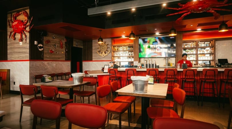 Interior of a seafood restaurant with red chairs, wooden tables, a bar, and crab decorations.