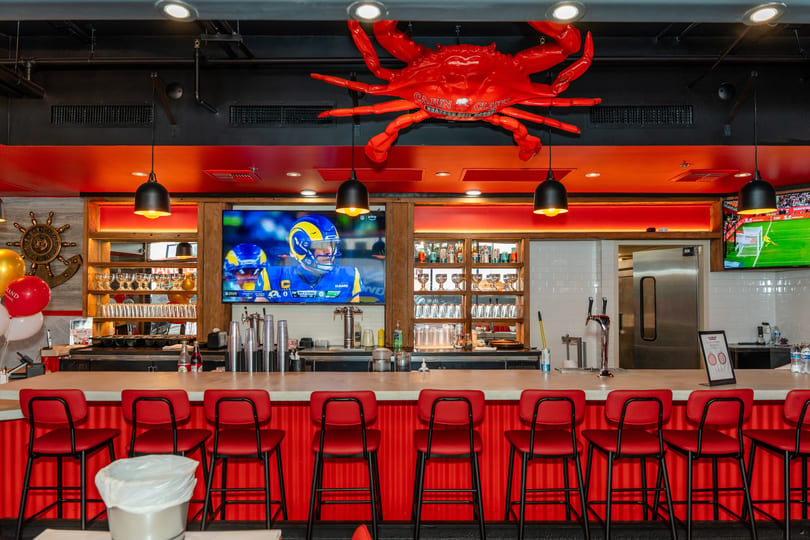 A vibrant bar with red stools, a large TV, and a giant red crab decoration on the ceiling.