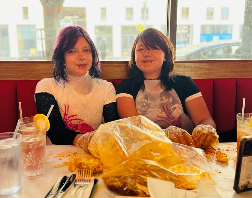 Two young women in plastic bibs and gloves, smiling after eating a messy seafood boil.