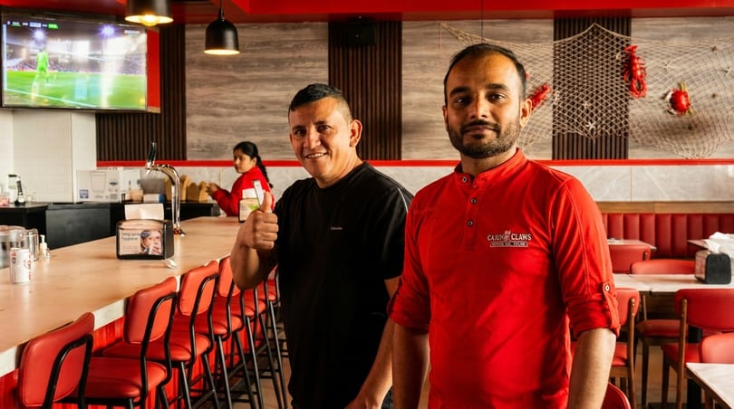 Two men smiling in a Cajun Claws restaurant, one giving a thumbs up at the bar.