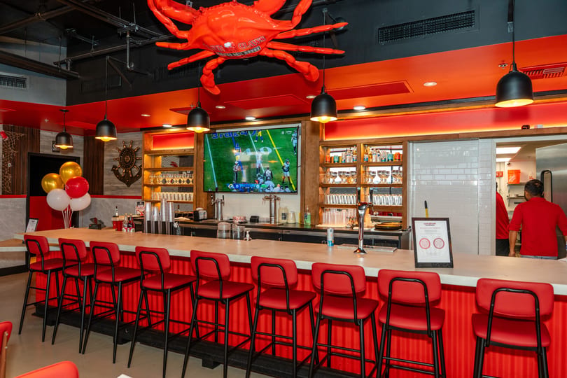 A vibrant bar with red stools, a large TV showing football, and a giant red crab decoration overhead.
