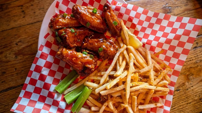 Sweet and sour chicken wings with french fries, celery sticks, and a lemon wedge on checkered paper.