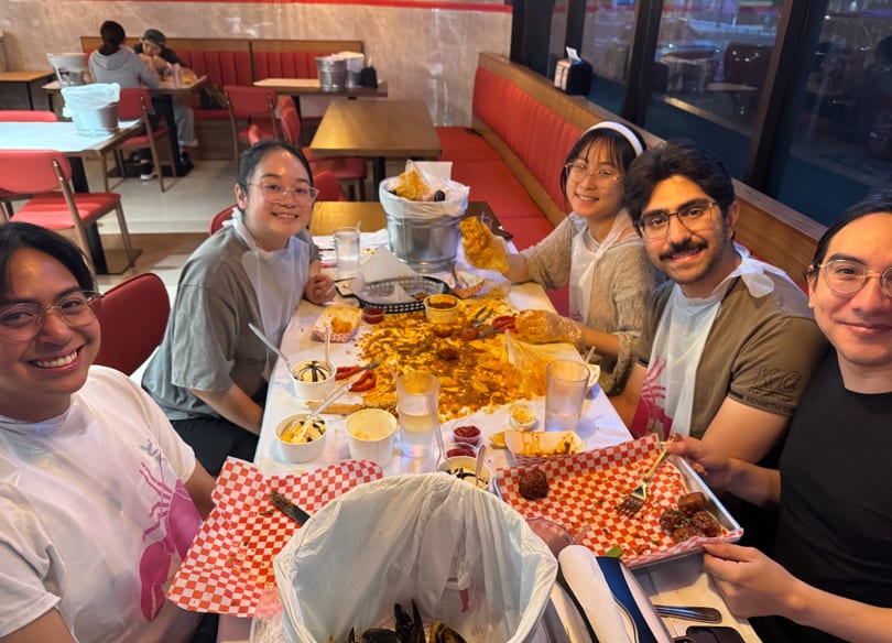 Five smiling friends wearing bibs and eating a messy seafood boil at a restaurant table.
