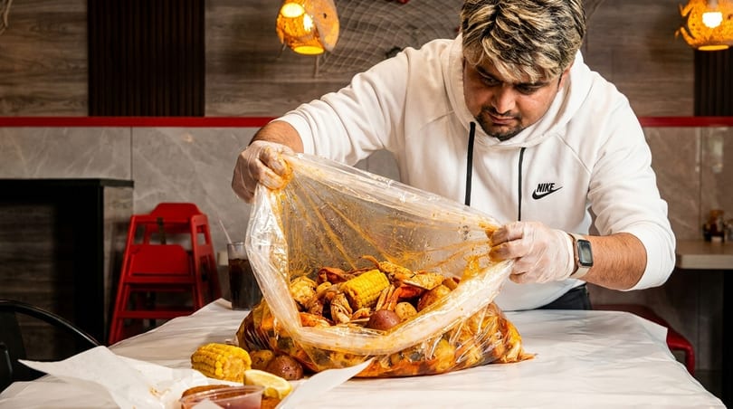 Man in gloves opening a large plastic bag filled with seafood boil, corn, and potatoes.